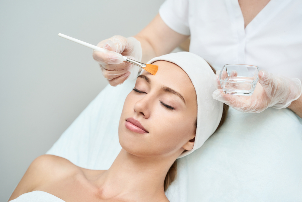 Skincare Specialist Performing A Professional Chemical Peel Treatment On A Woman Lying With Eyes Closed, Demonstrating A Facial Exfoliation Procedure For Rejuvenated Skin.